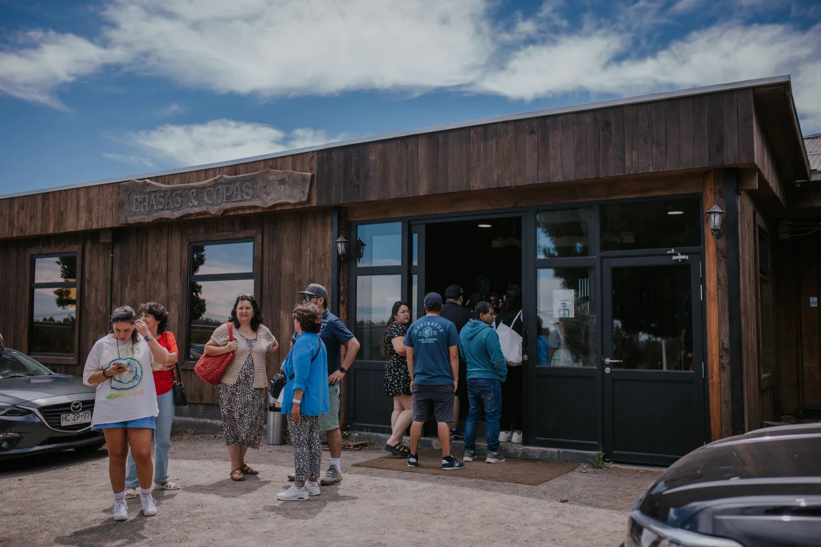 Fachada de madera del restaurante Brasas y Copas con cartel y clientes haciendo fila en la entrada bajo cielo azul