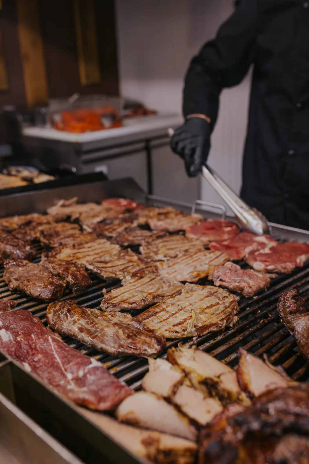 Variedad de cortes de carne con marcas de parrilla cocinándose mientras un parrillero los voltea con pinzas