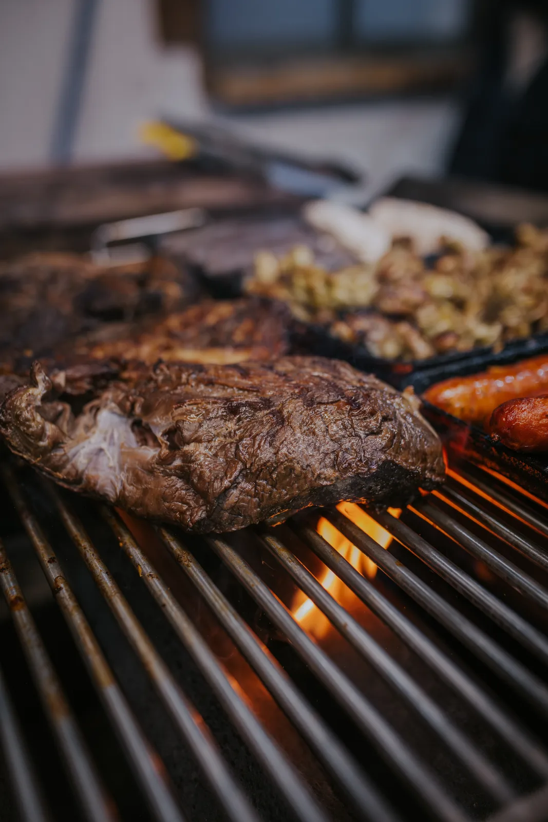 Corte de carne dorándose sobre parrilla encendida con llamas, junto a longaniza y mariscos